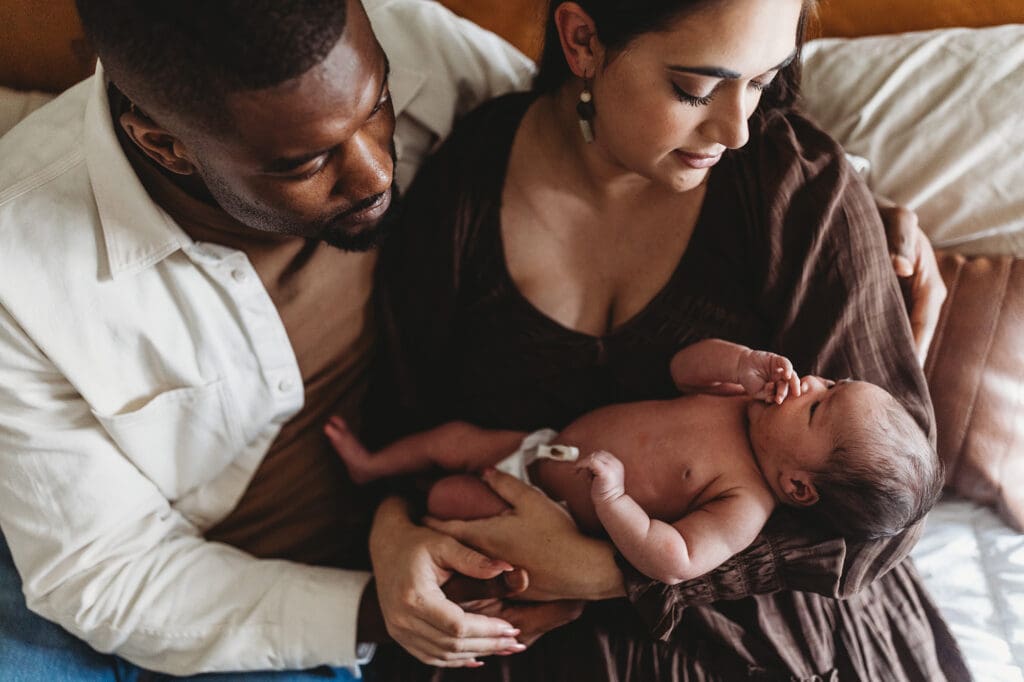 Atlanta newborn photographer capturing new parents gazing at their baby in a moody, neutral-toned in-home lifestyle session with rich brown colors