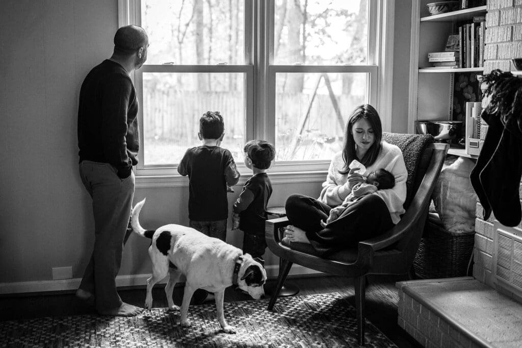 Black and white in-home newborn photo in Marietta with mom feeding baby, dad and sons looking out window, family dog in background