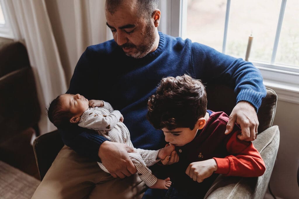 Atlanta in-home newborn photography with dad sitting and holding baby while older brother kisses newborn’s foot