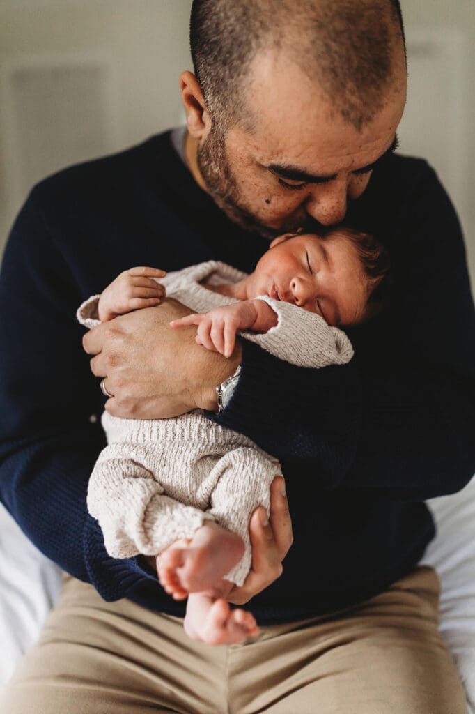 Marietta newborn photographer capturing dad holding baby against his chest facing camera, dad kissing newborn during in-home session