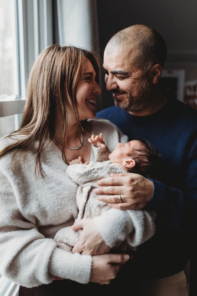 Marietta newborn photographer capturing mom and dad holding their baby by window light smiling at each other during in-home session