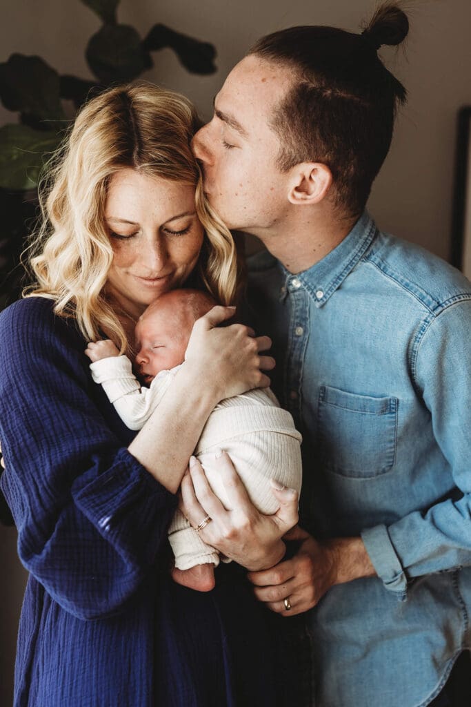 Mom and dad by window light, dad kissing mom while she snuggles their newborn during a Decatur newborn photography session.