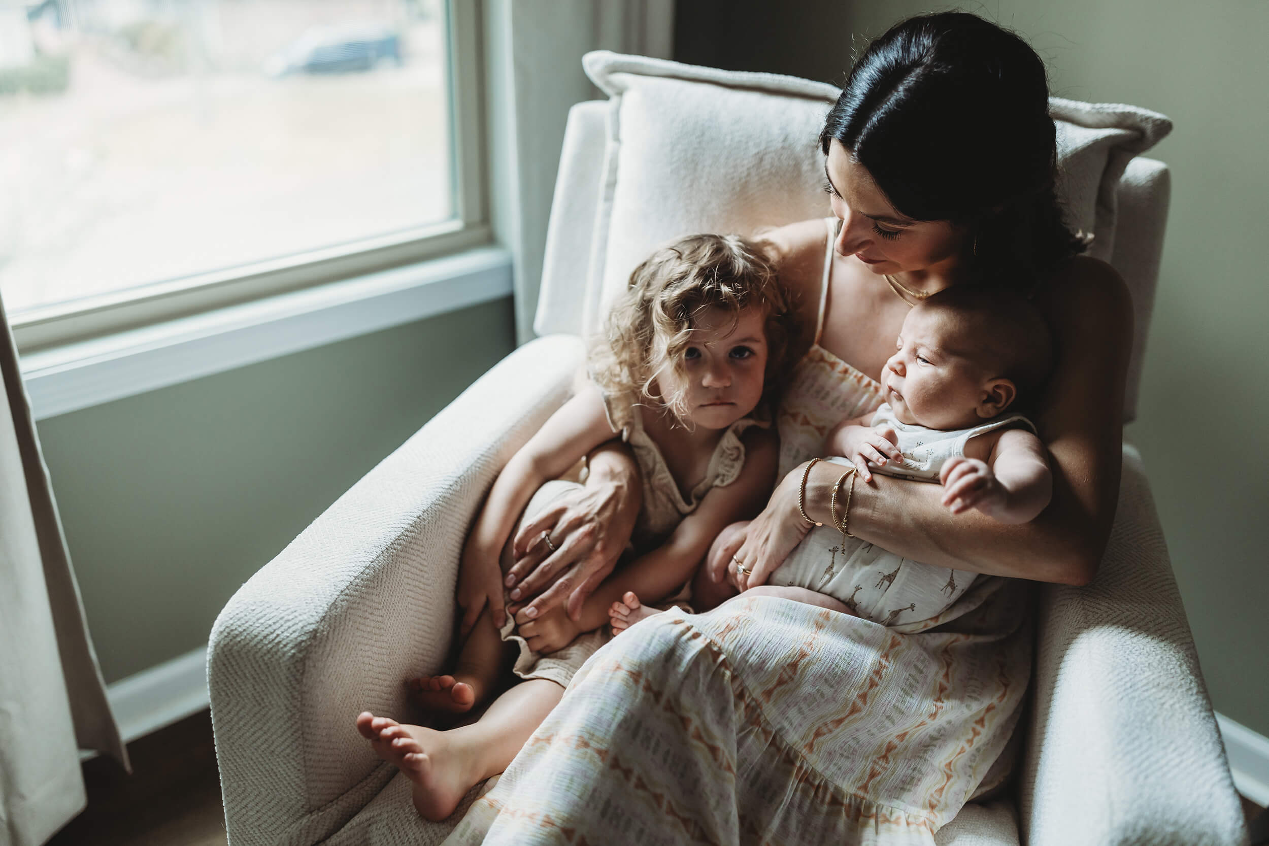 Mother with newborn baby and big sister by window light during a Sandy Springs session.