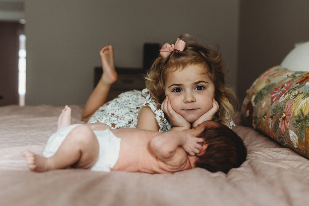 Big sister in a floral dress with a bow lying beside her newborn baby sister on the bed during an Atlanta lifestyle newborn photography session.