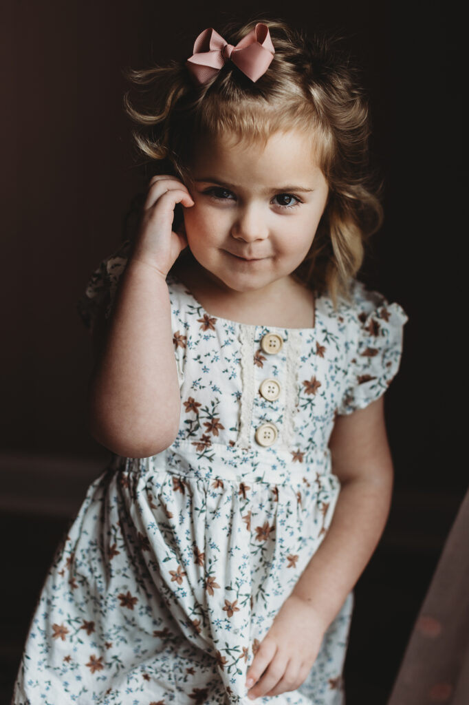 Older sister with soft curls and a pink bow standing by a window in a floral dress during a newborn photoshoot in Atlanta.