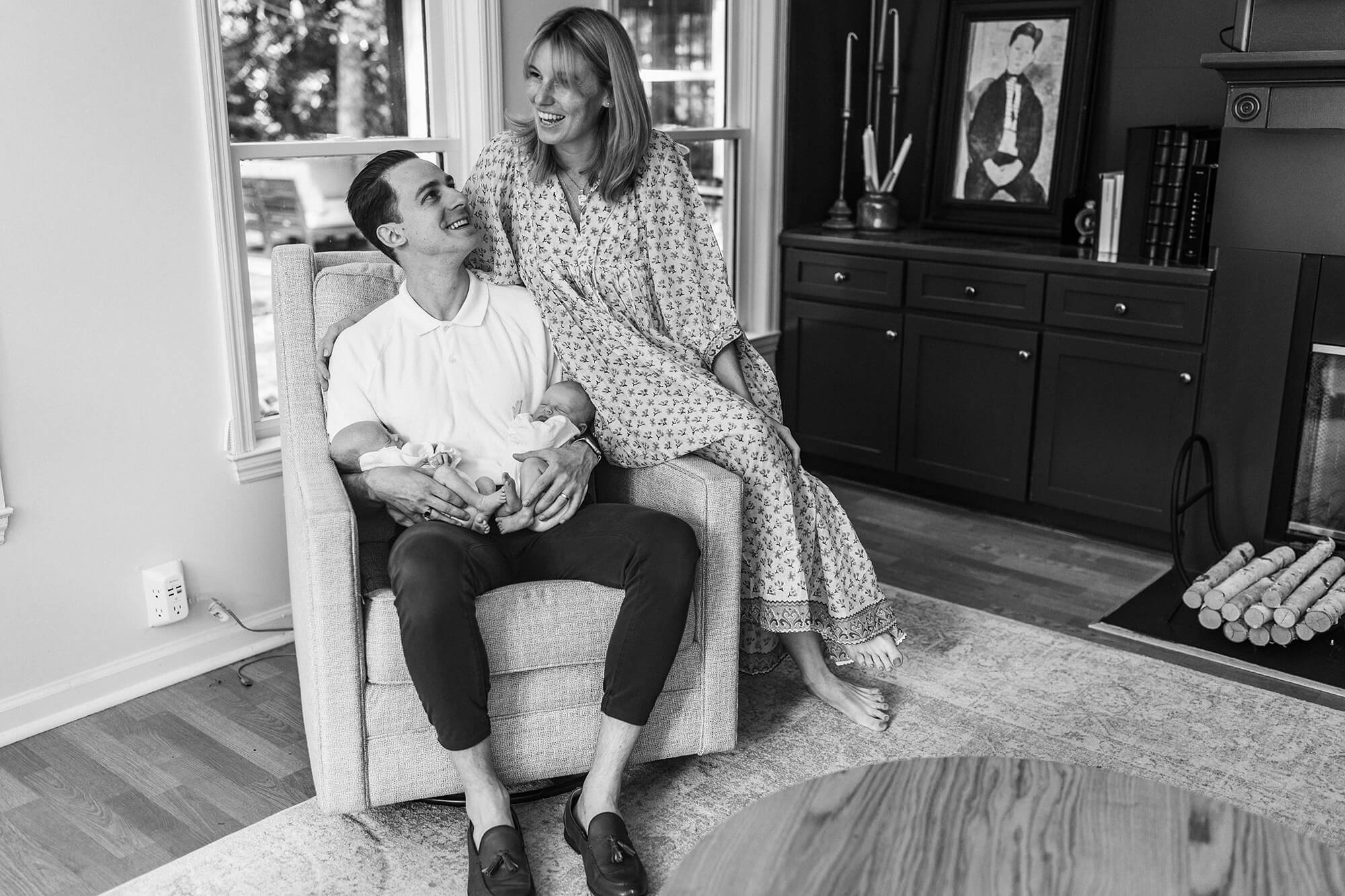 Black and white portrait of dad sitting in an armchair holding his newborn twin girls while mom sits on the arm of the chair laughing during an Atlanta newborn photography session.