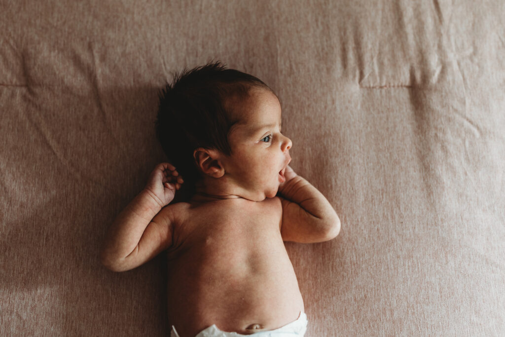 Newborn baby girl lying on a bed near a window with natural light illuminating her face as she yawns during an Atlanta newborn photography session.