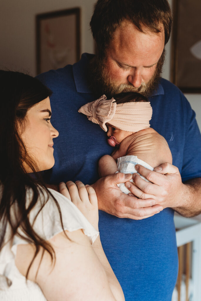Dad holding newborn baby by a window while mom smiles over his shoulder during a lifestyle session with an Atlanta newborn photographer.