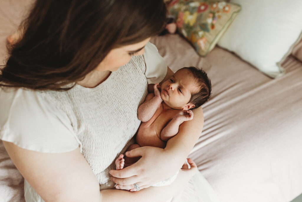 Mom in a cream dress sitting on a bed holding her awake newborn baby girl during an Atlanta lifestyle newborn photography session.