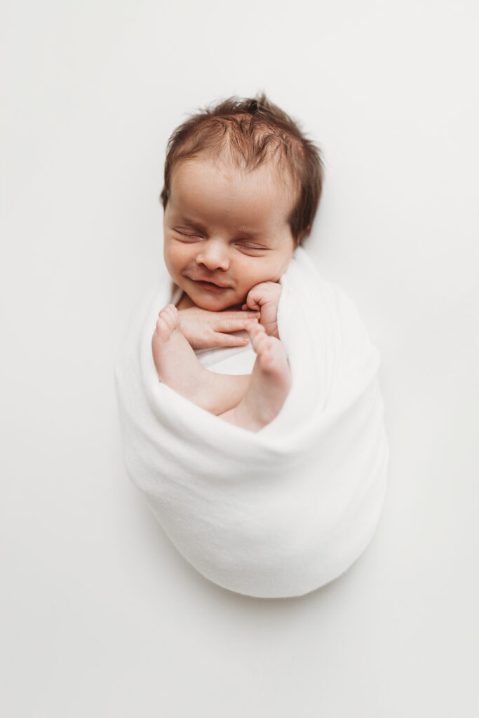 Sleeping newborn baby wrapped in white on a white backdrop smiling during a studio newborn photography session in Atlanta.