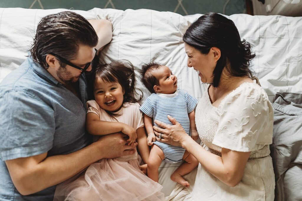 Atlanta newborn photographer capturing a candid in-home family moment with parents lying on the bed with their newborn baby boy and smiling toddler daughter in Decatur, Georgia, natural lifestyle newborn photography