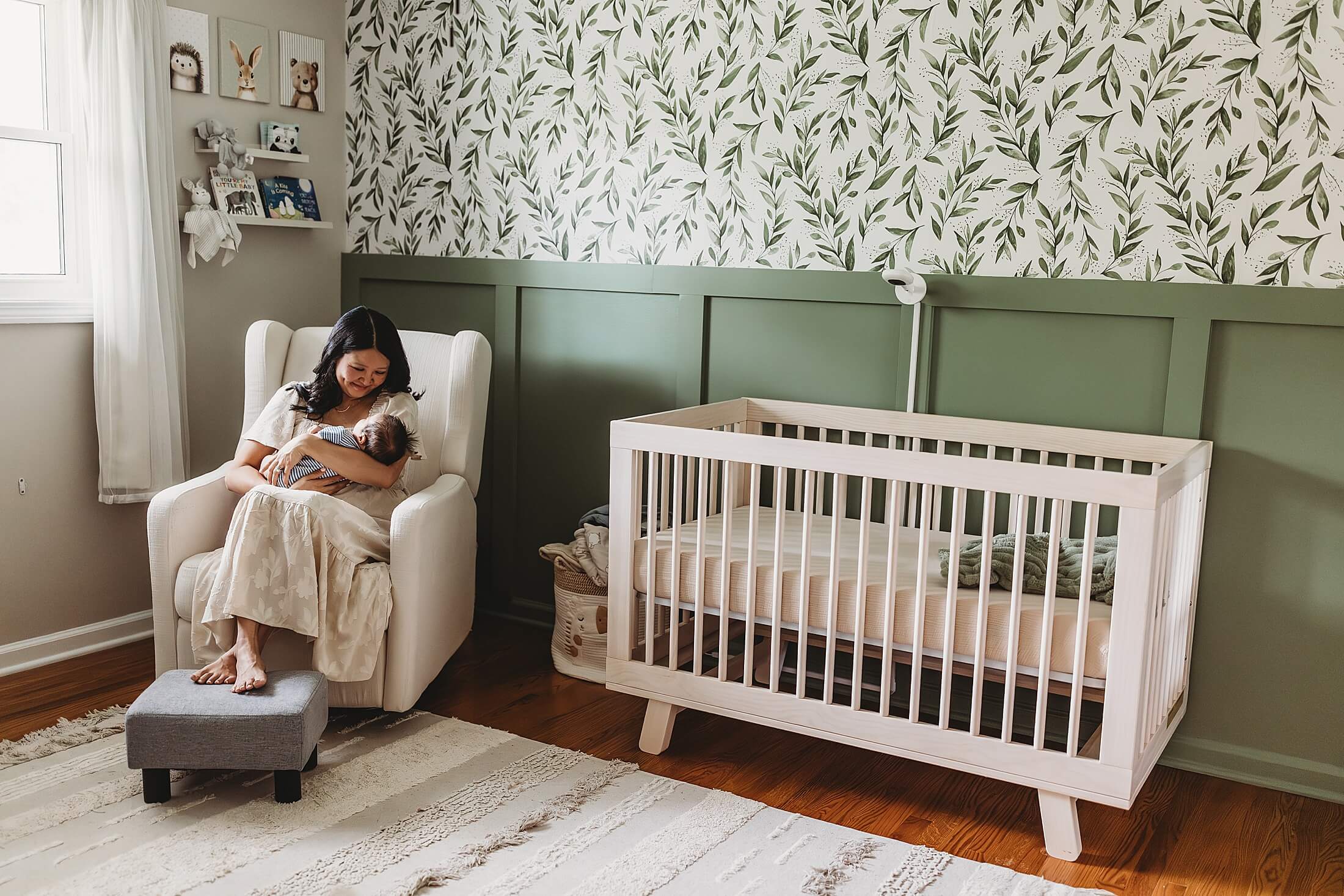 Atlanta lifestyle newborn photographer capturing a wide nursery view with sage green accent walls and mom sitting in an armchair with the baby boy, beautiful in-home newborn photography in Decatur, Georgia
