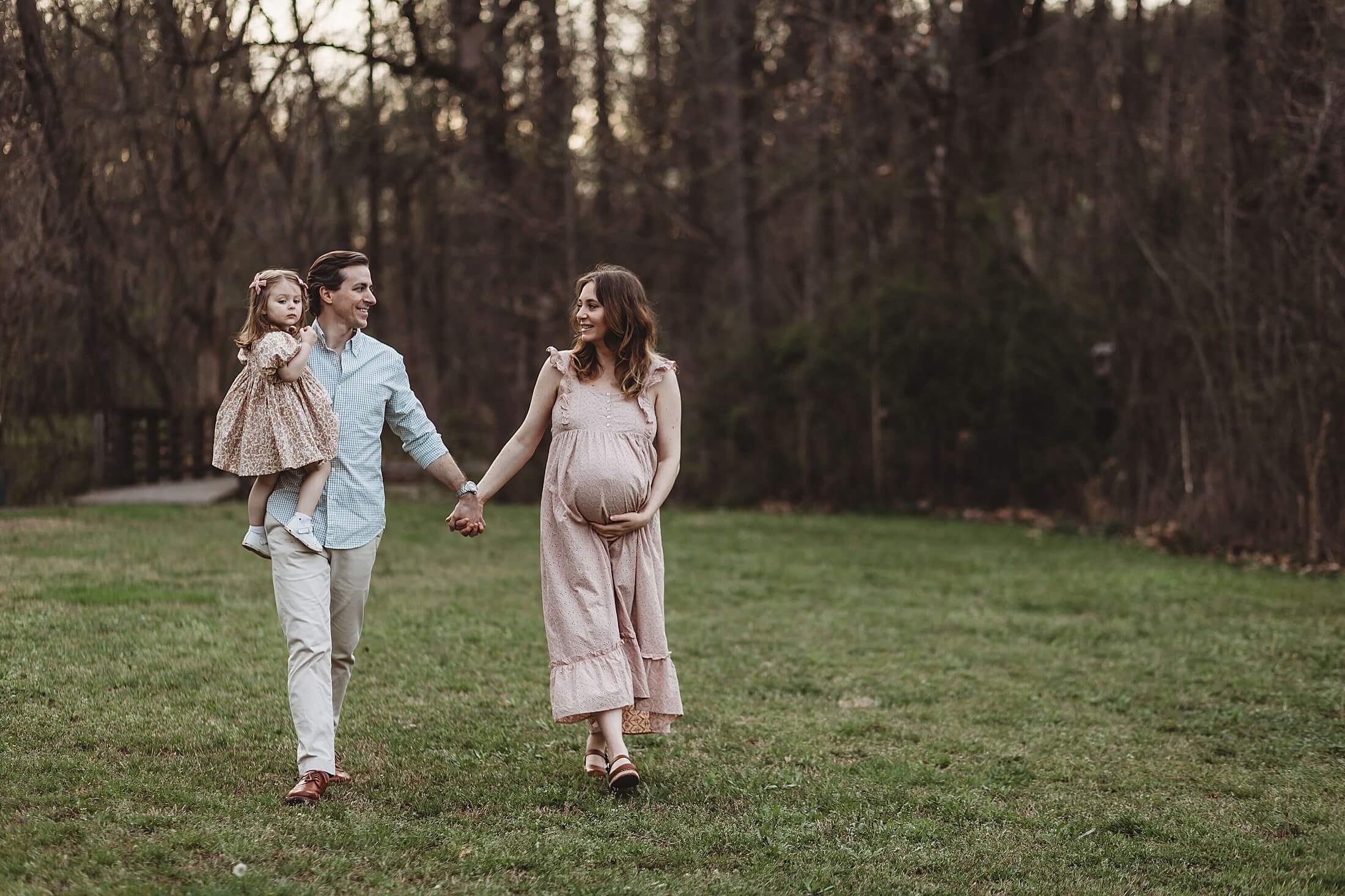 Family maternity session in Atlanta with parents walking hand in hand alongside their toddler during a candid outdoor pregnancy photo shoot