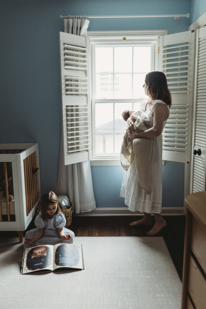 Mother holding her newborn by soft window light while toddler daughter sits on the floor reading a book, creating a warm and intimate lifestyle newborn photography moment in Atlanta