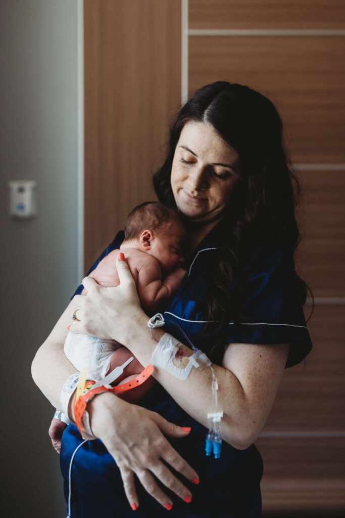 Mom wearing soft blue pajamas holds her newborn baby in only a diaper during a Fresh 48 session at Piedmont Hospital, both wearing hospital bracelets while sharing a cozy, intimate moment in the hospital room.