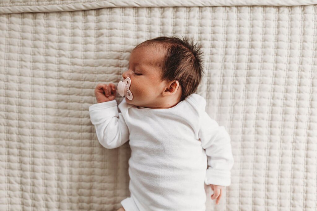 Newborn baby in a white onesie lying on a white bed during an Atlanta in-home newborn photography session.