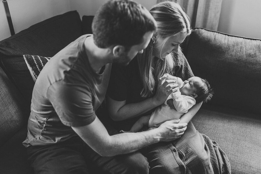 Black and white image of parents sitting together on a couch while mom feeds their newborn baby during an Atlanta in-home newborn session.