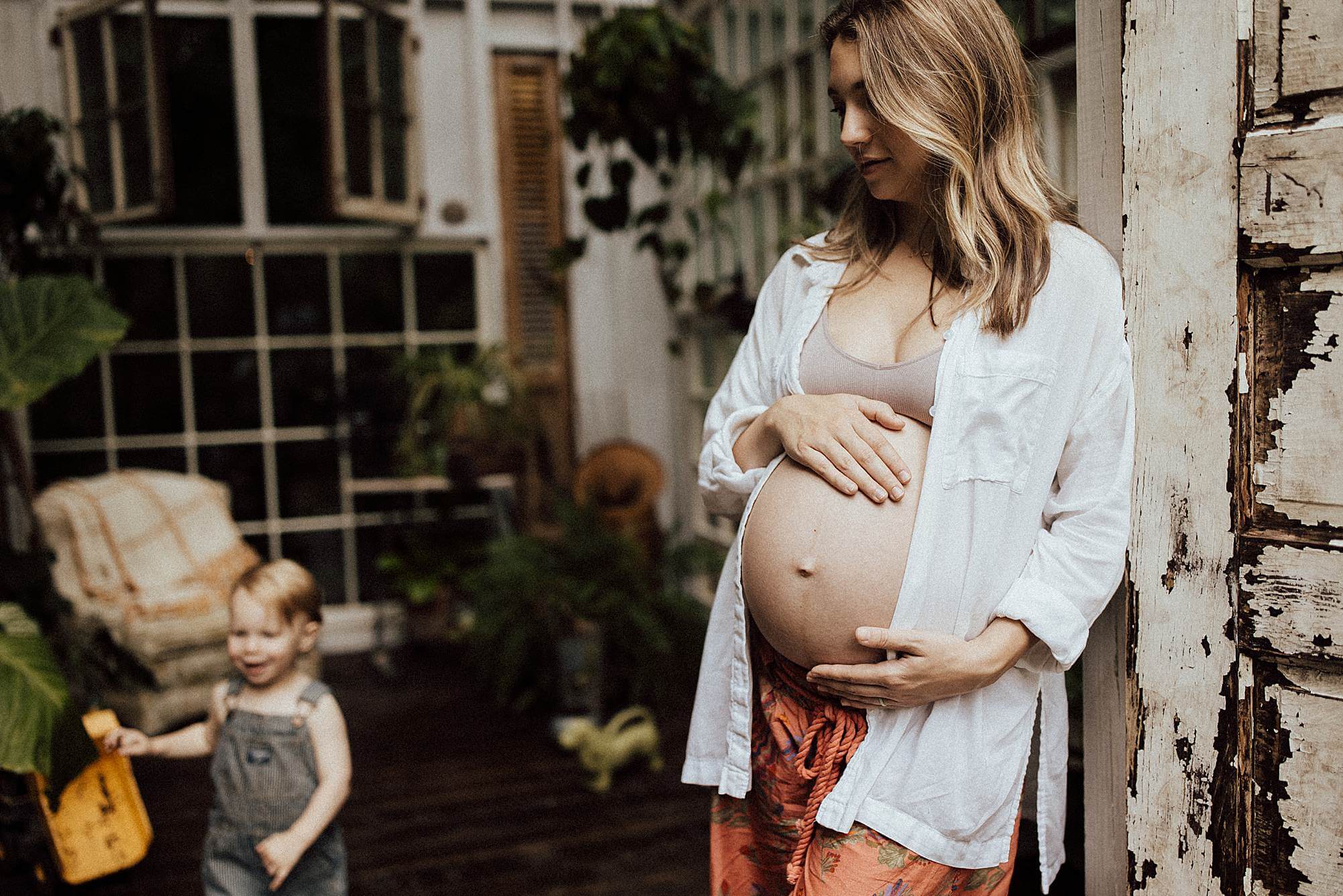 Expecting mother cradling her baby bump while spending time with her toddler in a North Atlanta greenhouse maternity session