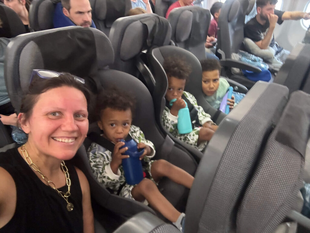 Children secured in car seats on a plane before takeoff during a family trip flying with a baby.